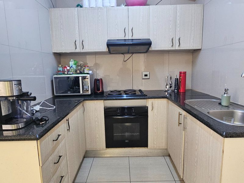 Compact kitchen featuring light wood-finish cabinetry, granite countertops, built-in oven, electric hob, extractor fan, and a stainless steel sink with tiled finishes.