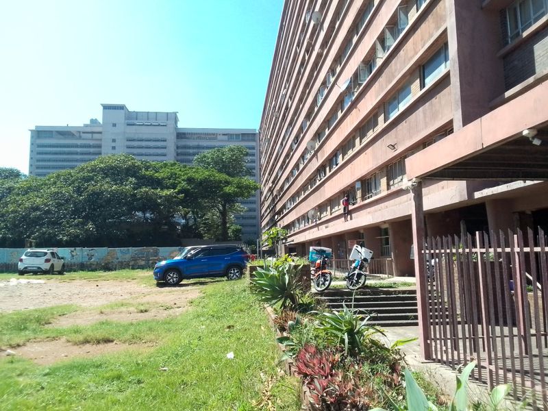 Exterior view of a multi-story apartment building featuring a concrete facade, metal security fencing, and an adjacent open parking area.