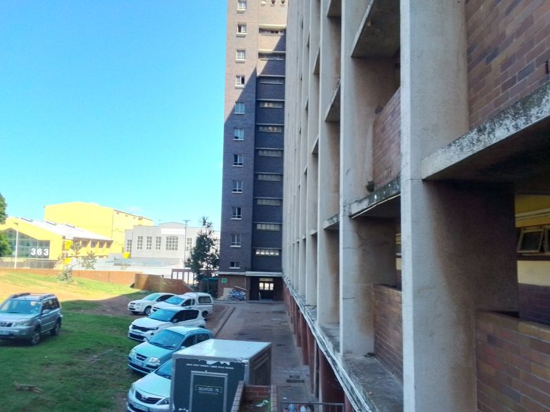 Exterior view of a multi-story apartment building featuring a brick facade and concrete balcony structures overlooking a parking area.
