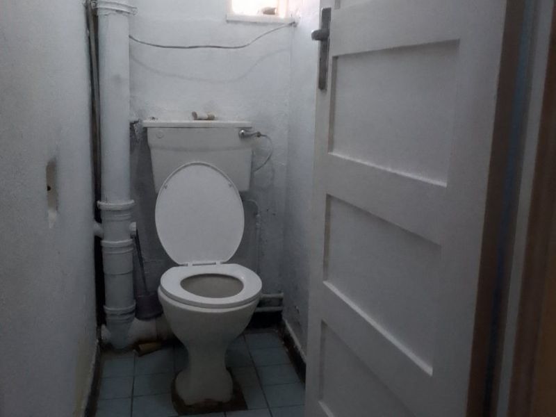 A small, dated bathroom featuring a standard toilet, exposed plumbing, and tiled flooring, requiring cosmetic updates and modernization.