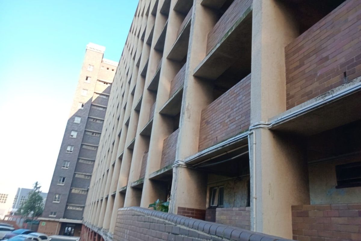 A low-angle exterior view of a multi-story apartment building featuring a repetitive brick and concrete facade with open balcony access.