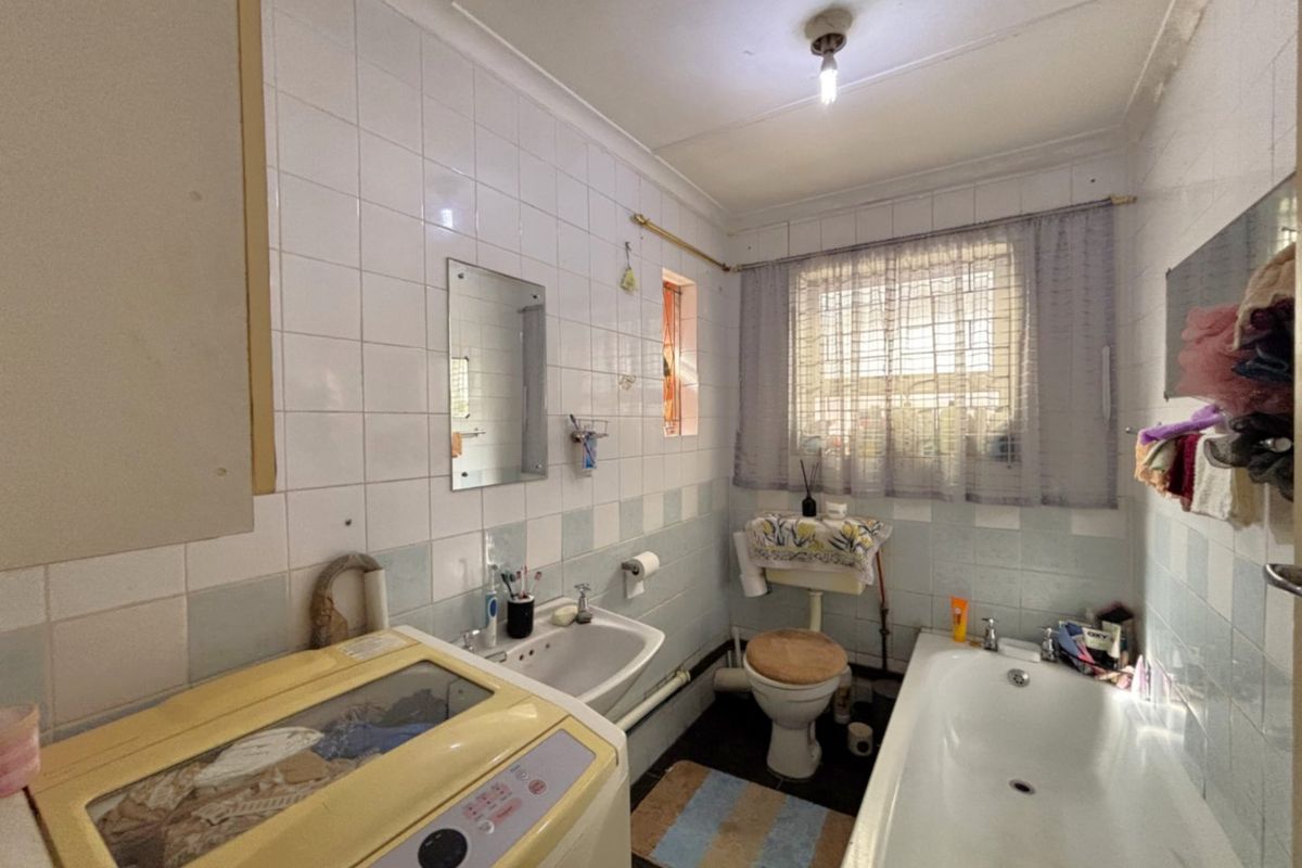 A dated bathroom featuring a bathtub, wall-mounted basin, and toilet, finished with ceramic wall tiles and a window with security bars.