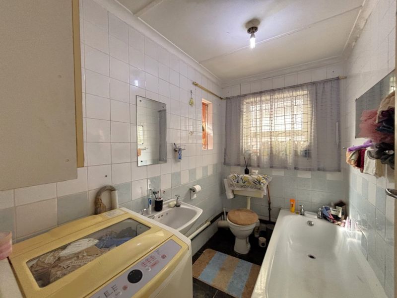 A dated bathroom featuring a bathtub, wall-mounted basin, and toilet, finished with ceramic wall tiles and a window with security bars.