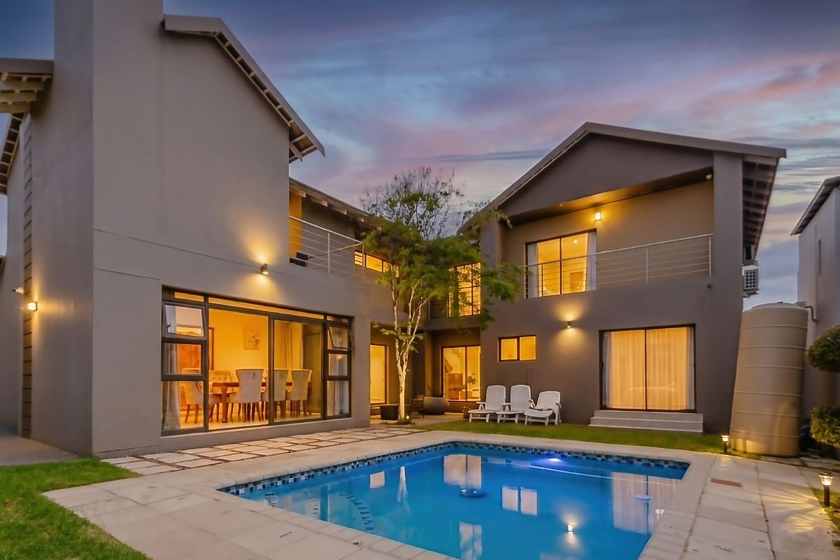 Rear view of a modern two-story house featuring a swimming pool, paved patio area, upper-level balconies, and integrated exterior lighting.