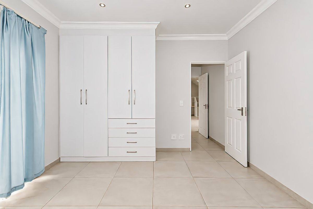 A bright, neutral bedroom featuring built-in white cupboards, tiled flooring, and recessed lighting, offering a clean and versatile space.