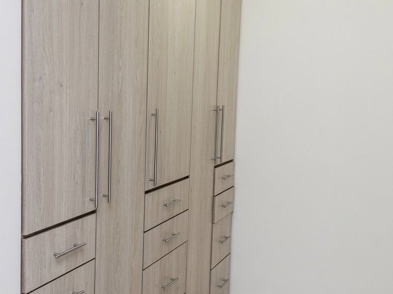 A clean, modern bedroom space featuring extensive light wood-grain built-in cabinetry with ample storage and neutral tiled flooring.