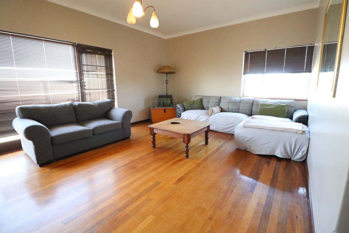 Spacious living room featuring polished hardwood flooring, neutral wall tones, and large windows fitted with venetian blinds.