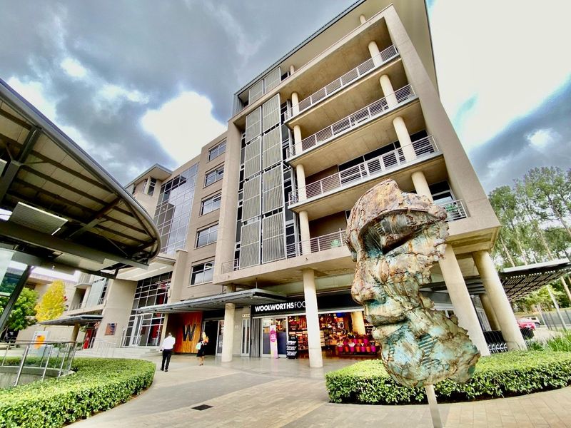 Modern multi-story commercial building exterior featuring a glass facade, balconies, and a paved pedestrian entrance area with landscaped hedging.
