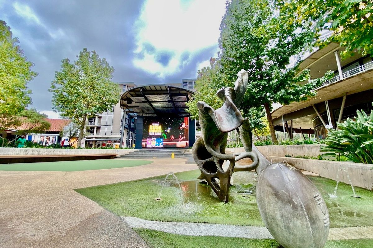 A well-maintained commercial outdoor plaza featuring a stage with a large LED screen, artistic sculpture, water feature, and landscaped garden areas.