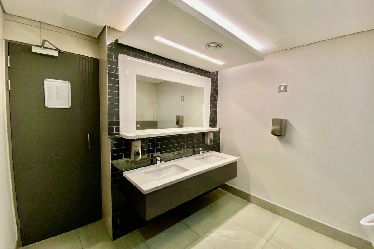 Modern commercial bathroom featuring a sleek double vanity with integrated mirror, subway tile backsplash, and recessed ceiling lighting.