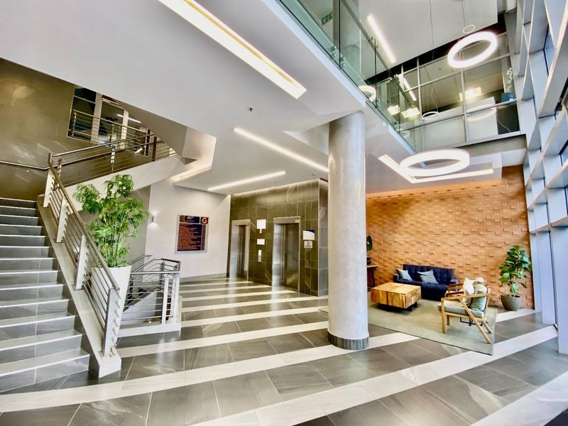 Modern commercial building lobby featuring a grand staircase, elevator access, high ceilings with recessed lighting, and a decorative textured feature wall.