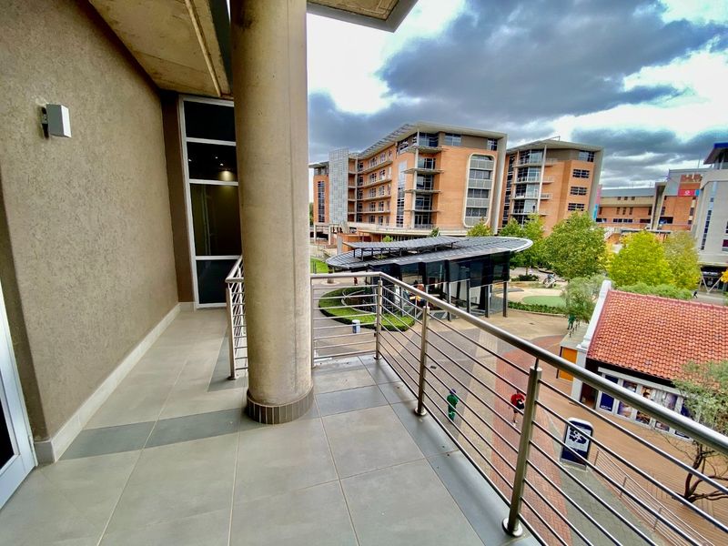 A spacious balcony featuring tiled flooring, a stainless steel balustrade, and a concrete support column, overlooking a commercial courtyard area.