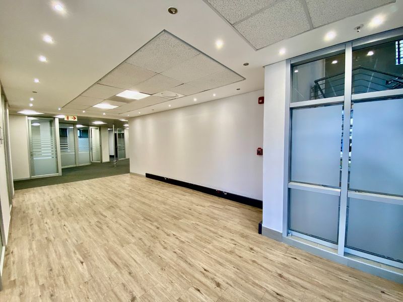 A bright, vacant commercial hallway featuring modern laminate flooring, suspended ceilings with recessed lighting, and glass partition walls.