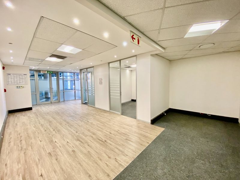 A bright, vacant commercial office space featuring a mix of laminate and carpet flooring, glass partition walls, and a modern suspended ceiling with recessed lighting.