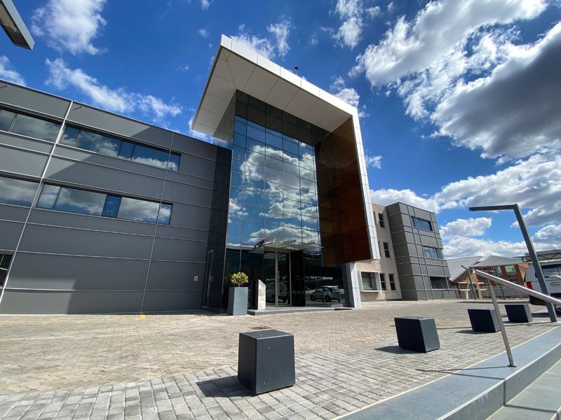 Modern commercial building facade featuring a prominent glass curtain wall entrance, metal cladding, and a paved forecourt area.