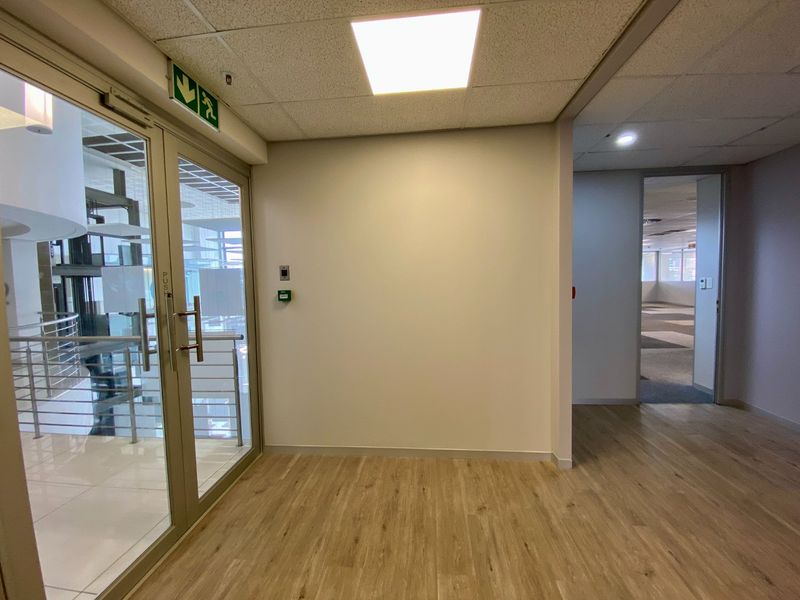 A clean, modern commercial hallway featuring wood-look flooring, recessed ceiling lighting, and a glass entrance door with integrated access control.