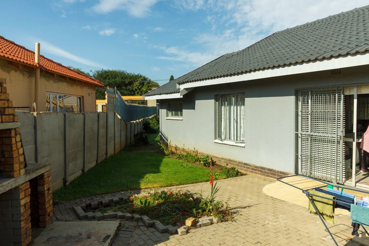 Rear exterior view featuring a paved patio area, built-in brick braai, and a small lawn enclosed by perimeter walling.
