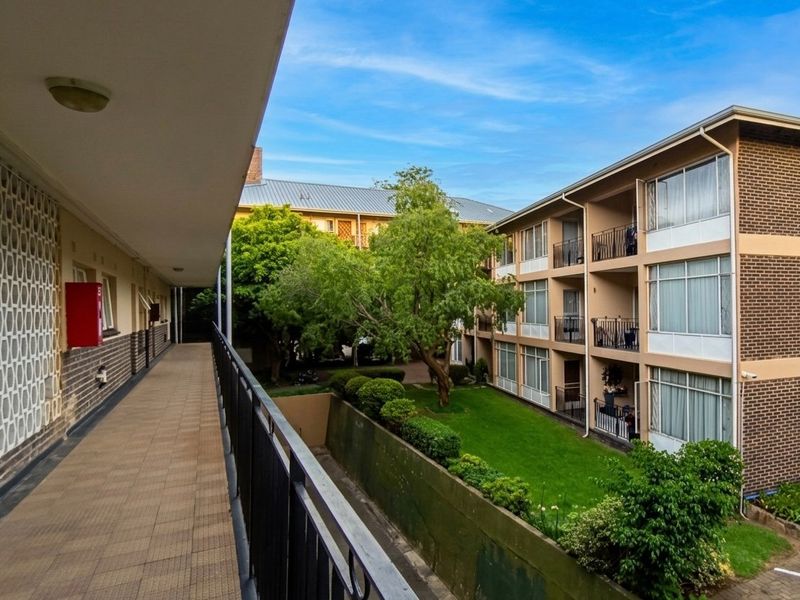 An elevated outdoor walkway featuring tiled flooring, a metal balustrade, and a decorative breeze block wall, overlooking the apartment complex courtyard and garden.