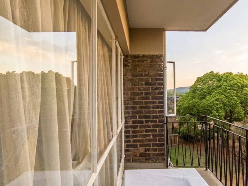 A private balcony featuring exposed brickwork, a wrought iron balustrade, and large glass windows overlooking the surrounding greenery.