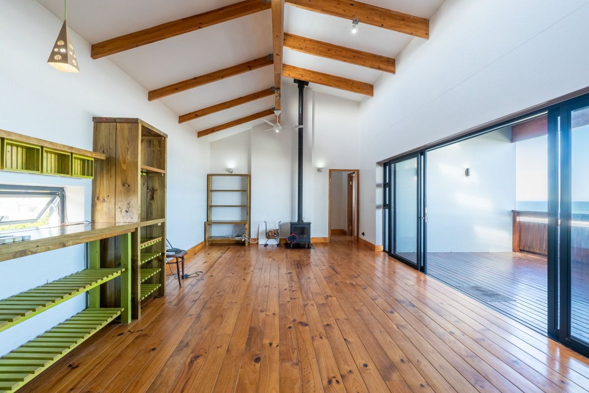 Spacious living area featuring vaulted ceilings with exposed timber beams, hardwood flooring, a wood-burning fireplace, and large sliding doors opening to a deck.