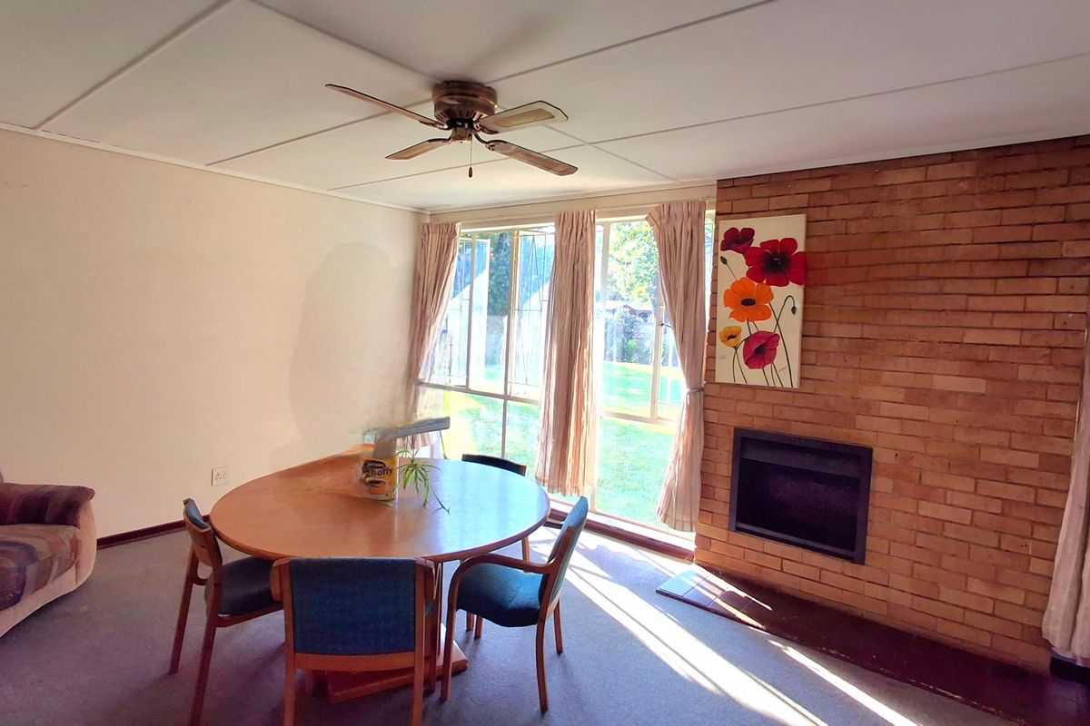 Living room featuring a prominent brick fireplace, ceiling fan, and large windows providing natural light and garden views.