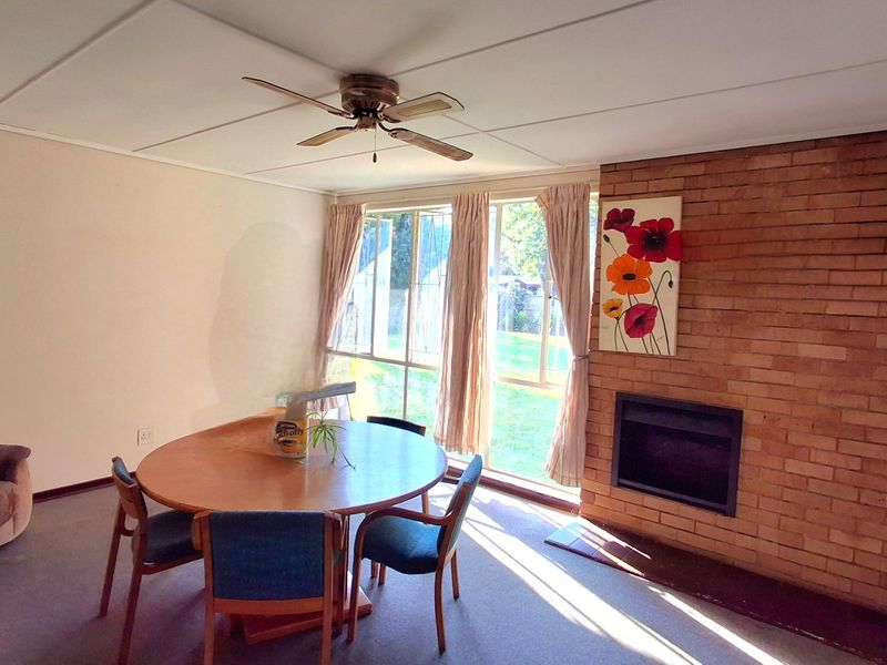 Living room featuring a prominent brick fireplace, ceiling fan, and large windows providing natural light and garden views.