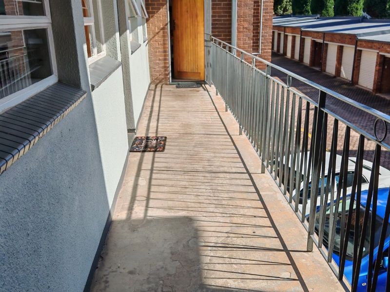An exterior balcony walkway featuring a metal balustrade, brick wall detailing, and a timber entrance door, showing signs of wear on the concrete floor.