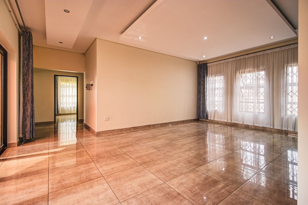 A spacious, empty living room featuring polished tiled flooring, recessed ceiling details with downlights, and large windows fitted with security gates.
