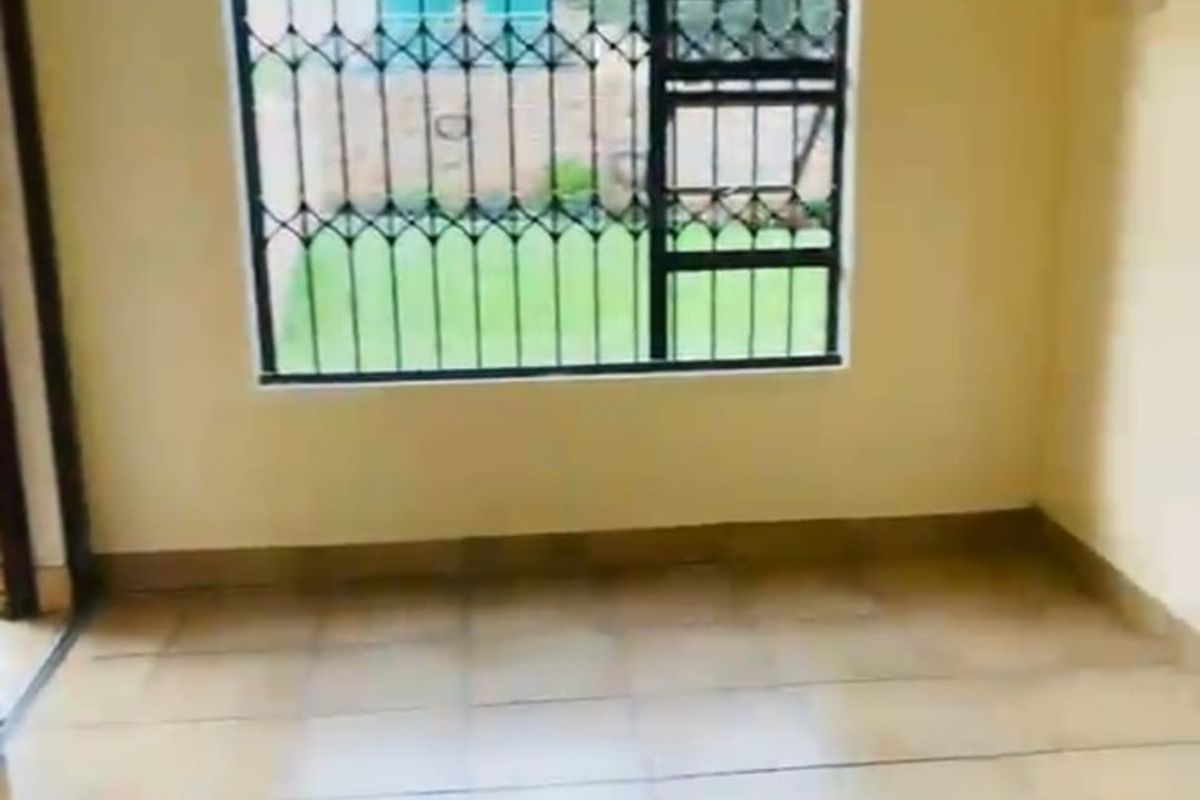 An empty living room featuring tiled flooring and a window fitted with security bars, offering a view of the garden.