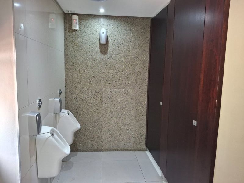 A clean, modern commercial restroom featuring wall-mounted urinals, tiled surfaces, a stone-textured feature wall, and private partitioned cubicles.