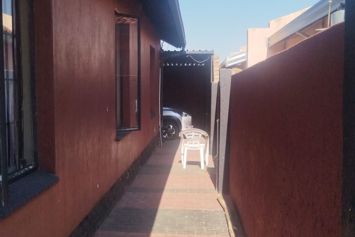 A narrow paved side walkway between the house exterior and a high boundary wall, leading towards a covered parking area.