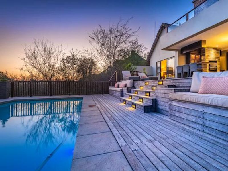A beautifully lit outdoor pool area featuring extensive timber decking, built-in tiered seating, and a modern fireplace, perfect for evening entertainment.