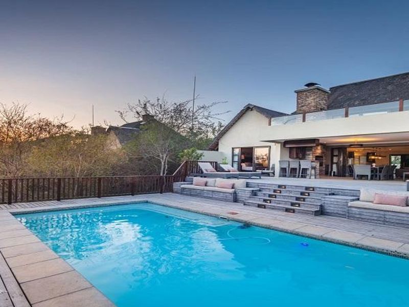 A stunning outdoor pool area featuring extensive timber decking, a built-in braai, and a glass-railed balcony overlooking the landscape at sunset.