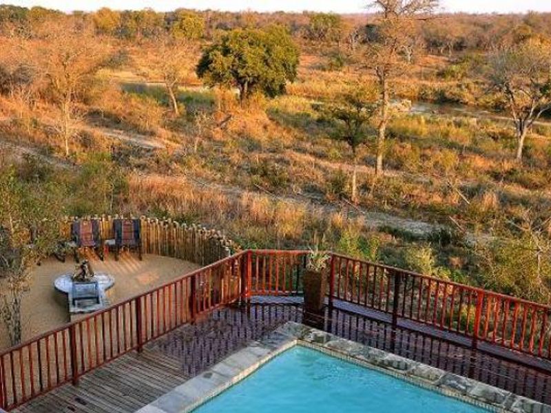 Elevated view of a swimming pool with a wooden deck and balustrade, overlooking a natural bushveld landscape with a boma fire pit area.