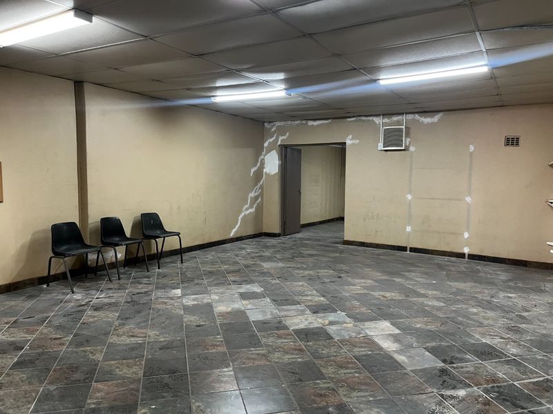 An empty commercial office space featuring tiled flooring, a suspended ceiling with fluorescent lighting, and a wall-mounted air conditioning unit.