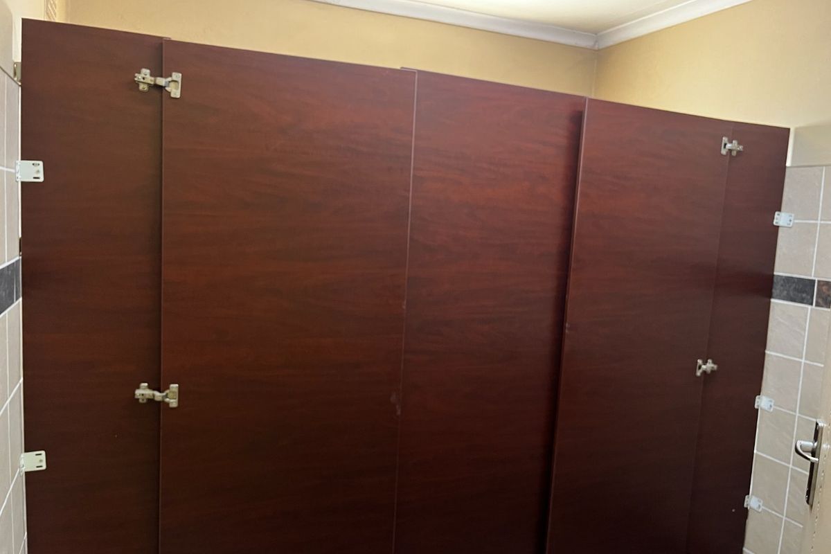 Commercial bathroom facility featuring dark wood-finish cubicle partitions and tiled wall surfaces, currently requiring maintenance on door hardware.