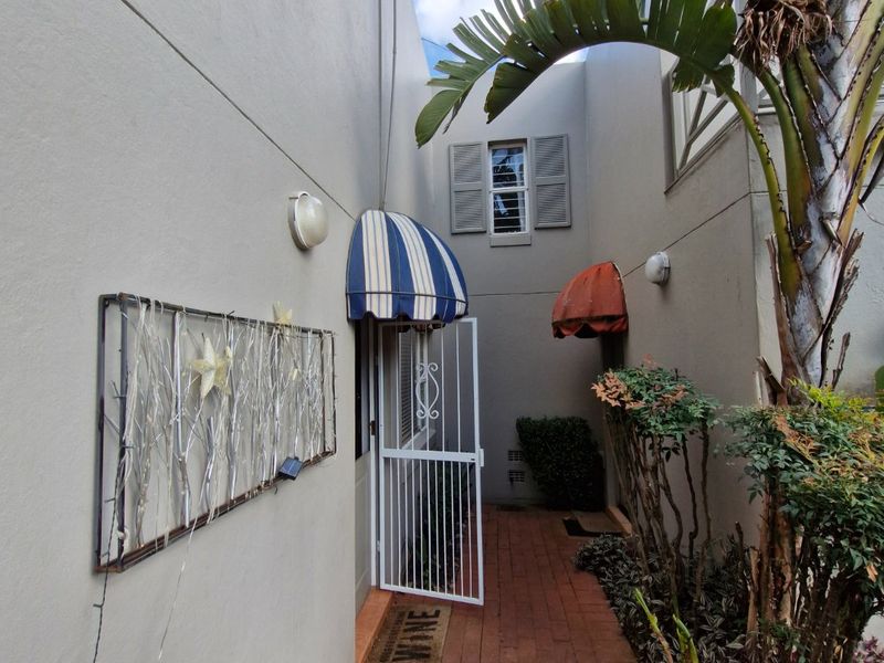 A narrow paved entrance walkway featuring a security gate, striped fabric awnings, and established garden greenery along the townhouse exterior wall.