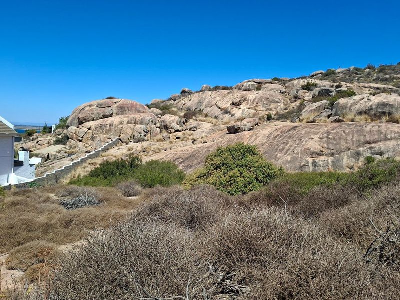 A view of a vacant residential plot featuring natural rocky terrain, native coastal vegetation, and existing concrete stairs leading up the slope.