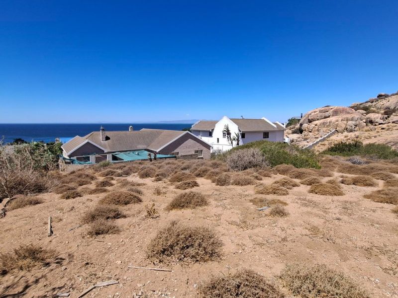 A view of a vacant residential plot featuring natural rocky terrain and coastal vegetation, with neighboring properties and the ocean in the background.