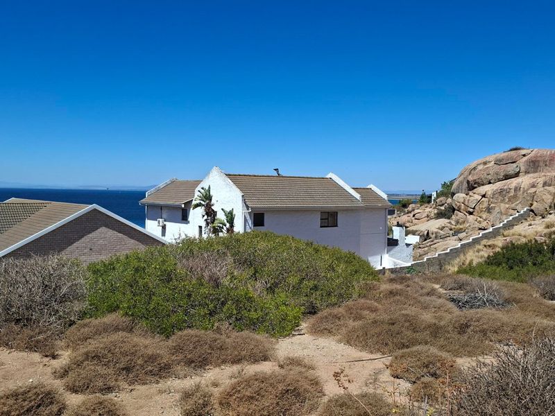 A view of the rear elevation of a coastal property featuring a tiled roof, white exterior walls, and a stone staircase set against a rocky landscape.