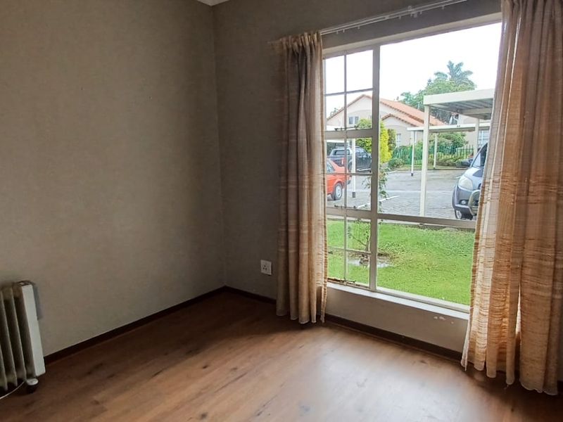 A bright, empty bedroom featuring laminate flooring, a built-in wardrobe, and a large window overlooking the complex exterior.