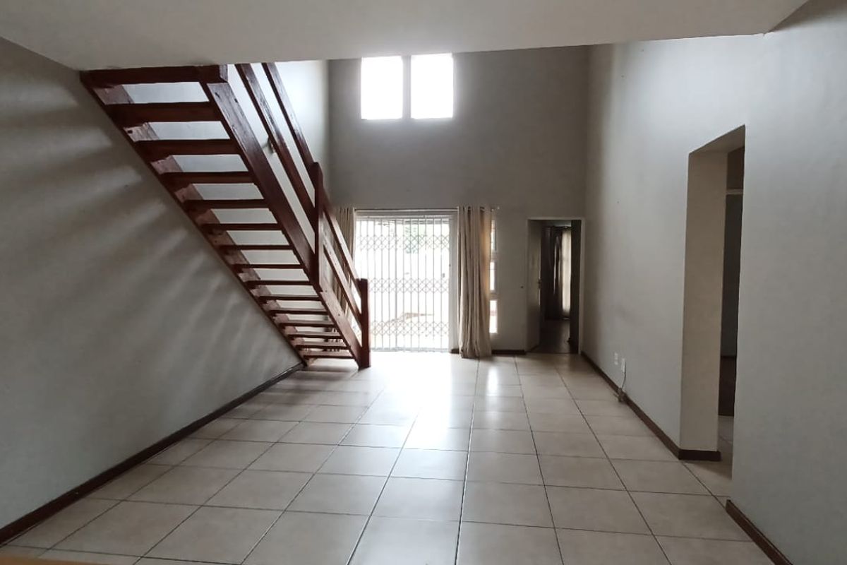 Spacious open-plan living area featuring tiled flooring, a wooden staircase, and double-volume ceilings with access to an outdoor area through a security gate.