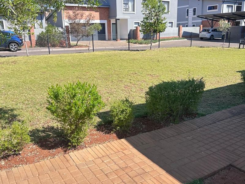 A view of a well-maintained rear garden featuring a paved walkway, manicured lawn, and perimeter fencing, overlooking neighboring townhouse properties.