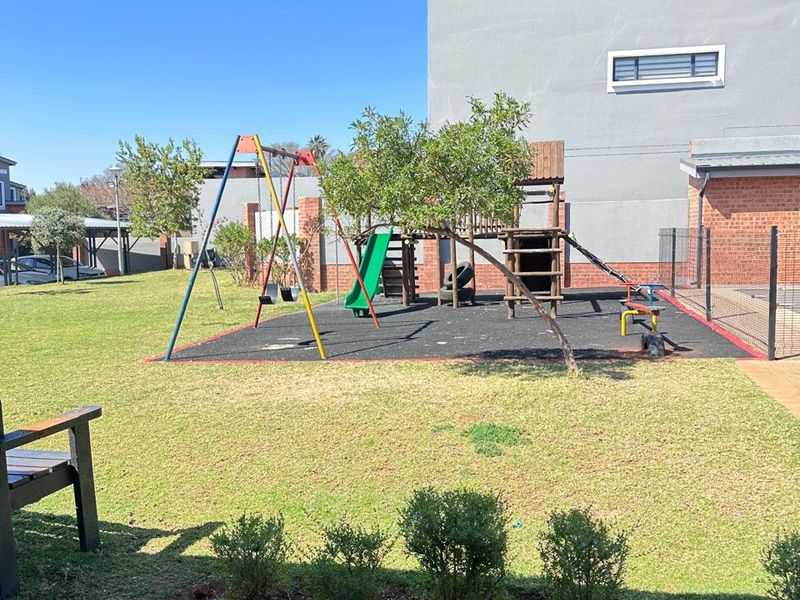 A well-maintained communal garden area featuring a paved patio, manicured lawn, and a children's play structure within a secure fenced perimeter.