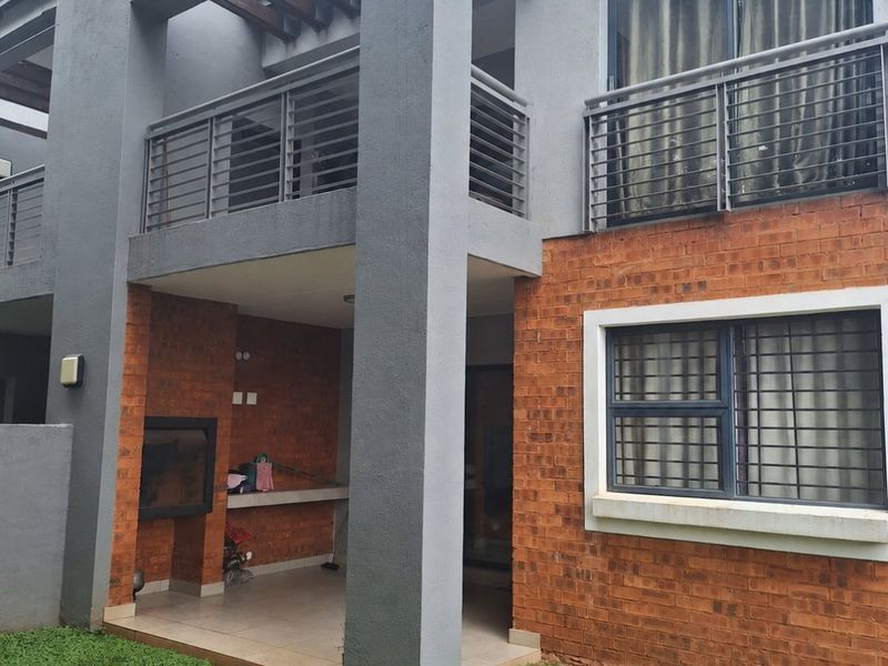 Rear view of a townhouse featuring a covered patio with a built-in braai, upper-level balcony, and a combination of brick and rendered facade finishes.