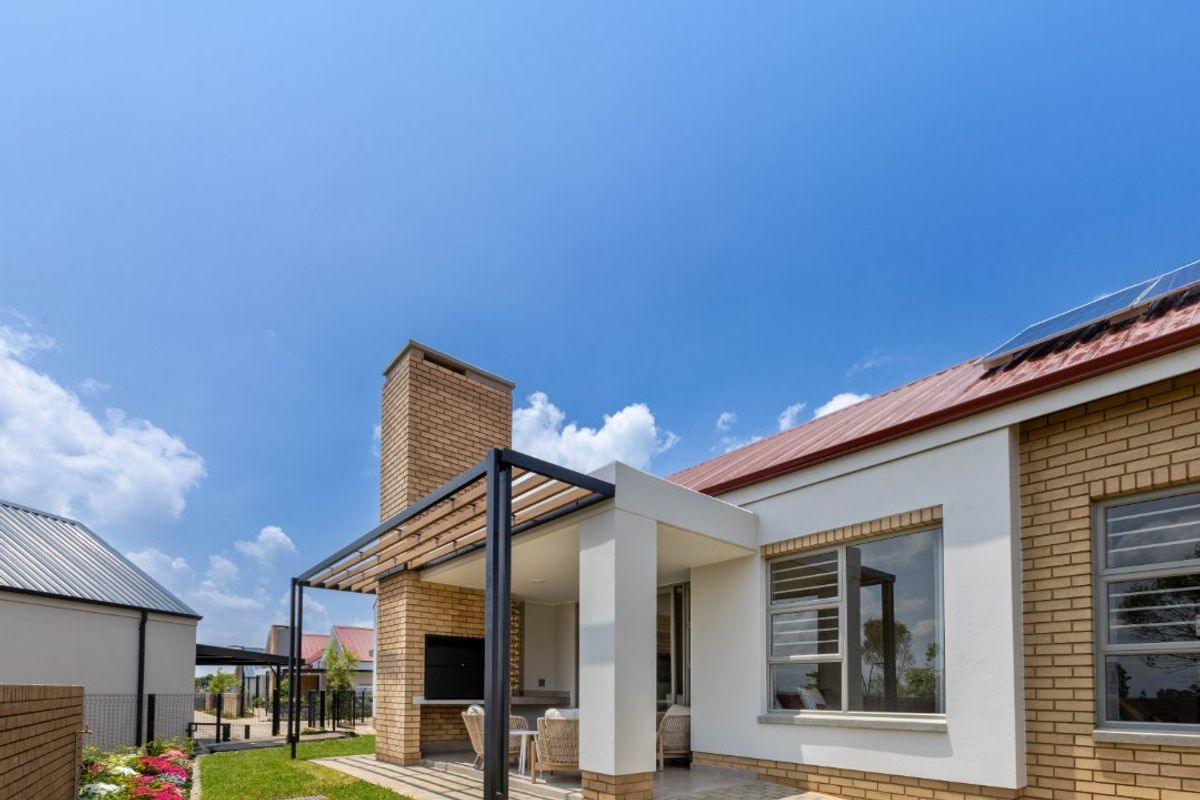 A well-maintained rear exterior featuring a covered patio with a built-in braai, brick chimney, landscaped garden, and solar panels on the roof.