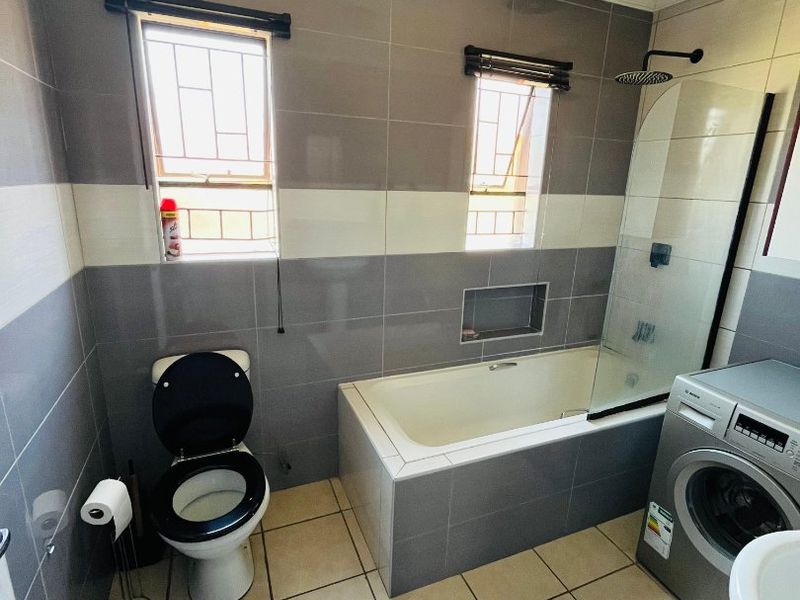 A modern bathroom featuring grey and white wall tiling, a built-in bathtub with a glass shower screen, and a toilet.