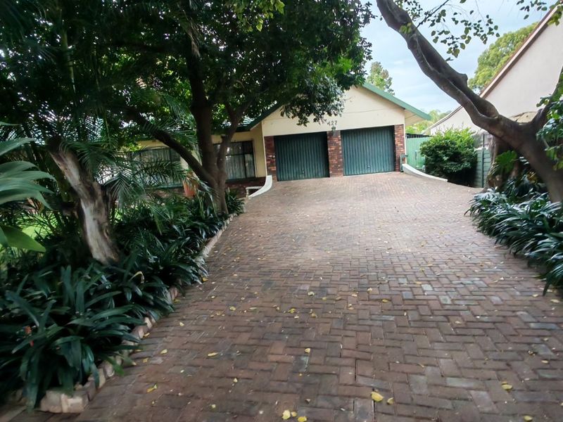 Front view of the property featuring a paved driveway leading to a double garage, surrounded by established trees and garden beds.