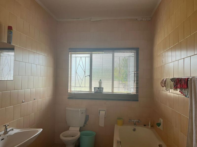 A dated bathroom featuring full-height ceramic wall tiles, a standard pedestal basin, a built-in bathtub, and a window with security bars.