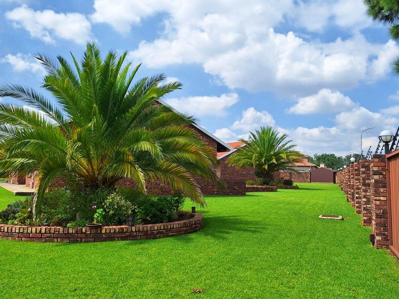 A well-maintained garden featuring a manicured lawn, established palm trees, and a secure brick boundary wall with electric fencing.